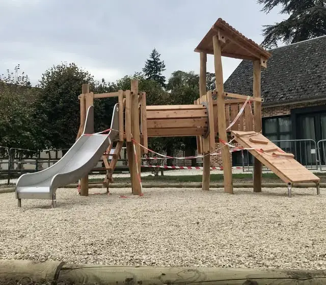 La nouvelle aire de jeux terminée sur la place du village : structure de jeu en bois avec toboggan métallique, devant l'école.